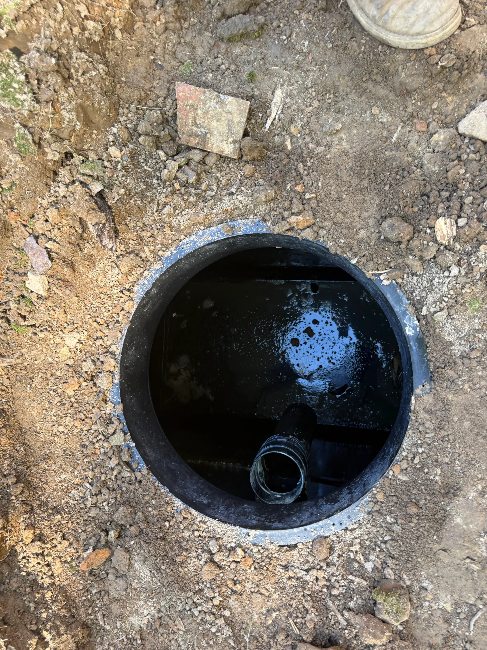 Open septic tank with water, concrete rim, and outlet pipe in excavated soil