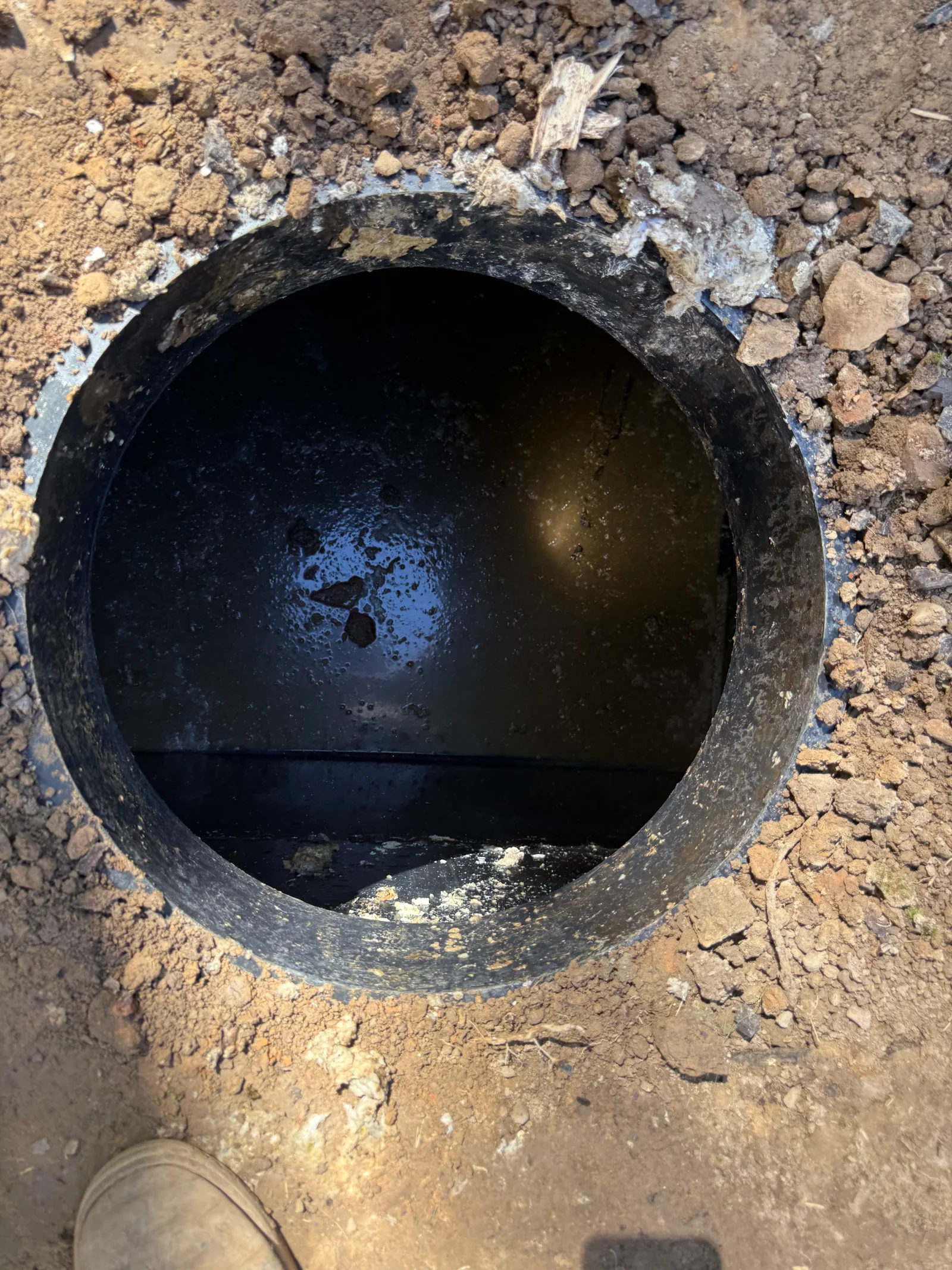 Open septic tank access with cast iron pipe, dark water, and surrounding excavated soil