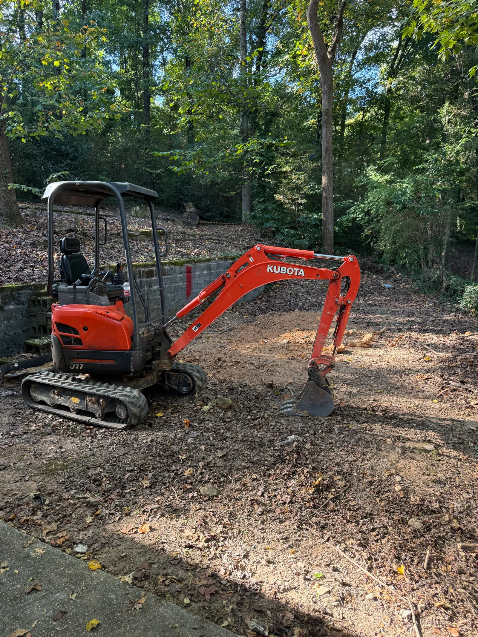 Red Kubota compact excavator with bucket on tracked base, excavating soil on wooded property